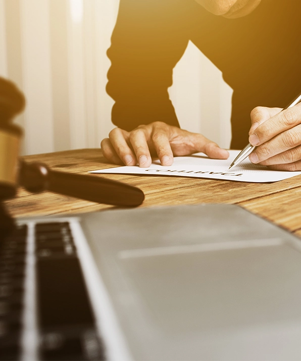 A person writing on a document labeled "Contract" beside a laptop and a gavel, suggesting legal work or agreements.
