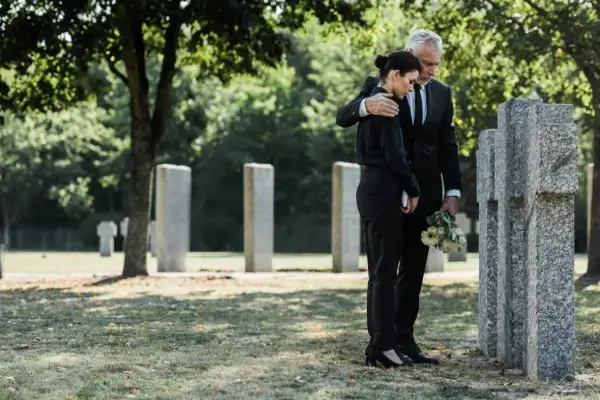 A grieving man in a suit comforts a woman in black clothing as they stand by gravestones in a cemetery