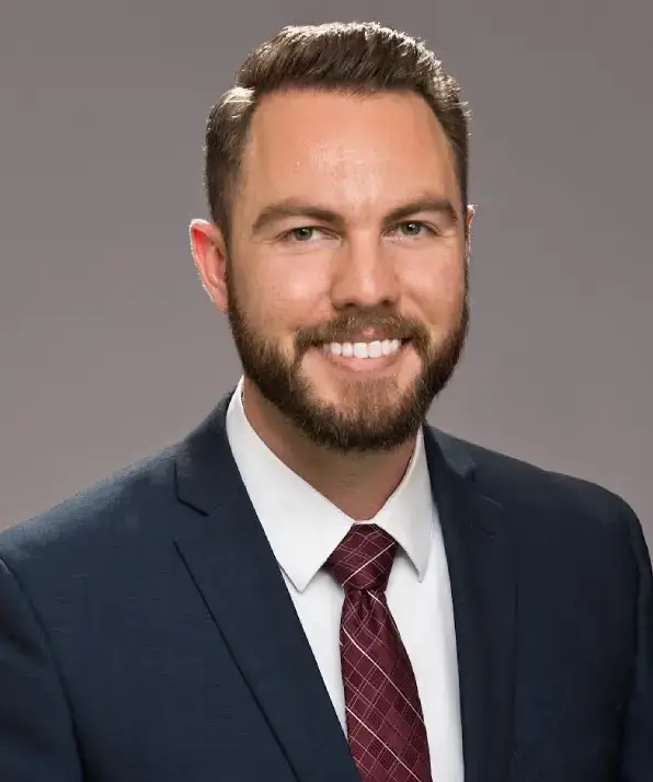 A man in a navy suit and maroon tie poses against a neutral gray background, exuding professionalism and confidence.