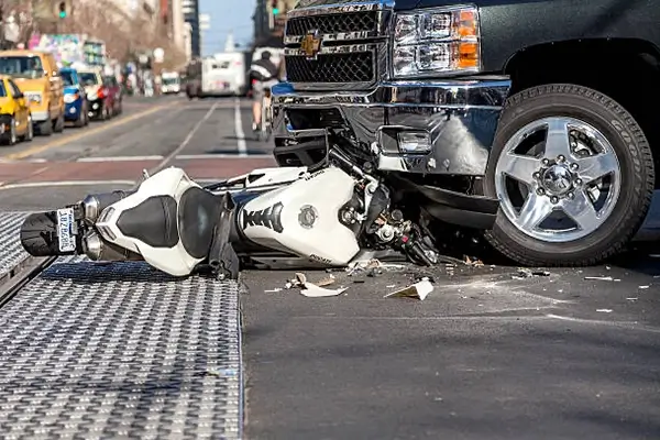 A white motorcycle lies on its side beneath the wheel of a black pickup truck, surrounded by debris on a busy urban street.